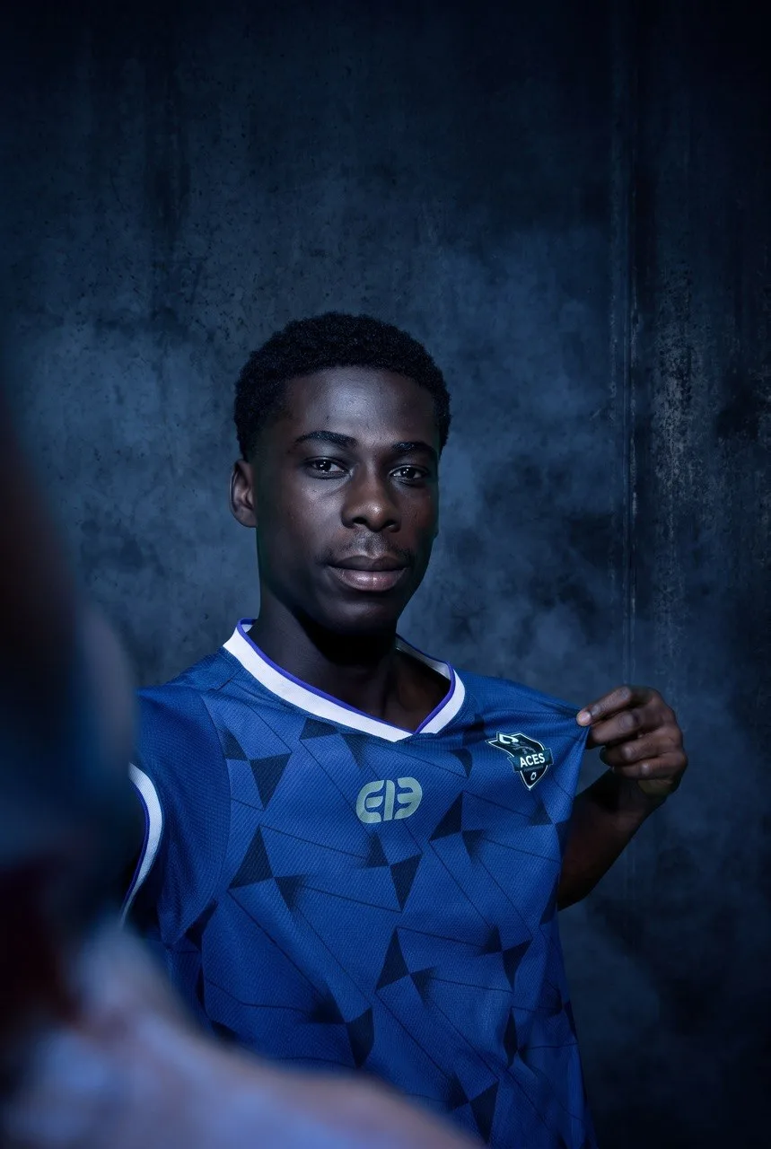 An Aces FC player in the club's blue home kit, lit against a dark studio backdrop.